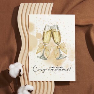 Könnte beinhalten: Eine Grußkarte mit zwei Sektgläsern, die anstoßen, jeweils mit einer goldenen Schleife verziert. Auf der Karte steht in eleganter Schrift "Congratulations!". Der Hintergrund ist ein weiches Beige mit goldenem Konfetti.