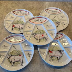 Op de afbeelding: Vijf witte keramische borden met verdeelde secties voor sauzen. Elk bord heeft een gedetailleerde illustratie van een koe met een slagersgids, gelabelde secties en tekst in het Frans. De borden zijn rond met een licht verhoogde rand.