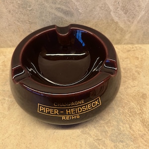 Op de afbeelding: Een donker bordeauxrode keramische asbak met een glanzende afwerking. De asbak heeft een afgeronde vorm met drie groeven om sigaretten op te leggen. De tekst "CHAMPAGNE PIPER-HEIDSIECK REIMS" is in gouden letters op de zijkant gedrukt.