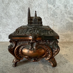 Pode incluir: Uma caixa de metal vintage adornada com um design detalhado. A tampa apresenta uma réplica em miniatura de um edifício de igreja.
