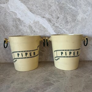 Op de afbeelding: Twee witte plastic champagne-emmers met groene handvatten en zwart-witte filmstrip-ontwerpen met daarop "Champagne Piper".