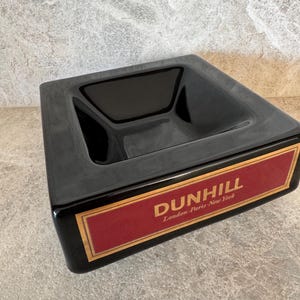Può includere: Un posacenere nero lucido, di forma quadrata con un interno incassato. Presenta un'etichetta rettangolare con il marchio "DUNHILL" in lettere dorate su sfondo rosso, con "London Paris New York" sotto.