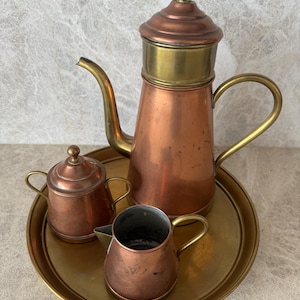 May include: A vintage copper and brass coffee set, including a tall coffee pot, a small creamer, and a sugar bowl with a lid. The set is displayed on a round brass tray. The coffee pot has a curved spout and a brass handle.
