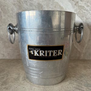 Op de afbeelding: Een zilverkleurige metalen ijsemmer met twee ringhandvatten. De emmer heeft een label met de tekst "KRITER" in een zwart en goud ontwerp. De emmer is cilindrisch met een licht uitlopende bovenkant.