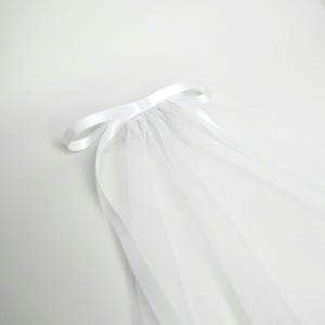 May include: A sheer white tulle veil featuring a smooth satin ribbon tied into a bow at the top. The delicate fabric gathers beneath the ribbon, creating soft, flowing folds that extend downwards in a translucent, layered arrangement.
