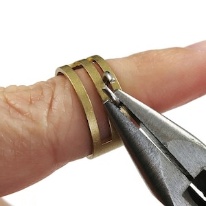 May include: A pair of pliers is being used to close a jump ring on a gold-toned ring. The ring is on a finger.