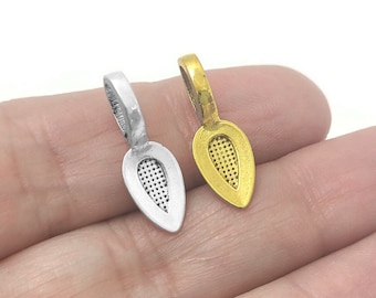 May include: Two teardrop-shaped pendant bails, one silver and one gold, with a textured design. The bails are shown on a hand.