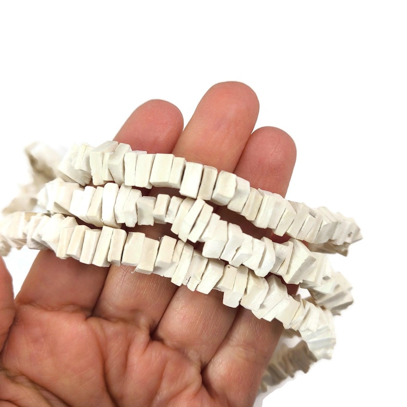 May include: Strands of off-white, rectangular beads are held in a hand against a white background. The beads have a slightly irregular shape and are strung together, creating a textured appearance. The beads are likely used for jewelry making.