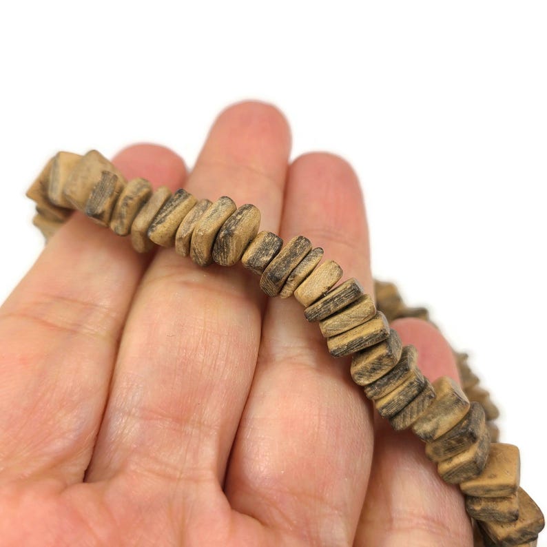 May include: A close-up of a beaded bracelet. The bracelet is made of small, square-shaped wooden beads in shades of brown and tan. The beads are strung together, and the bracelet is held in a person's hand against a white background.