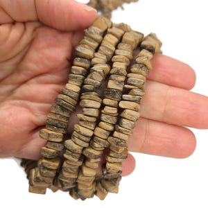May include: Strands of natural-toned wooden beads with a rectangular shape. The beads have a light brown color with darker brown accents. The beads are held in a hand against a white background. The beads are likely for jewelry making.