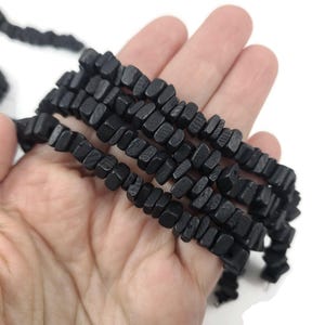 May include: Strands of black wooden beads with a rectangular shape. The beads are held in a hand against a white background. The beads have a textured surface and are strung together.