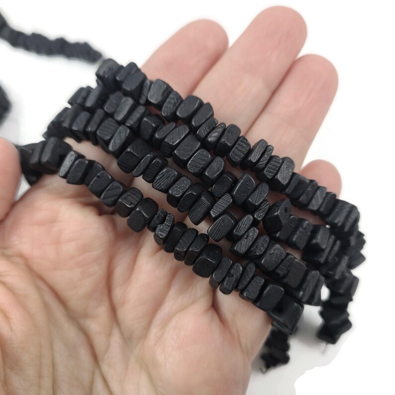 May include: Strands of black wooden beads with a rectangular shape. The beads are held in a hand against a white background. The beads have a textured surface and are strung together.