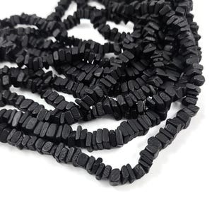 May include: Close-up of strands of black wooden beads. The beads are rectangular and have a slightly rough texture. The beads are strung together, creating a layered effect. The background is white.