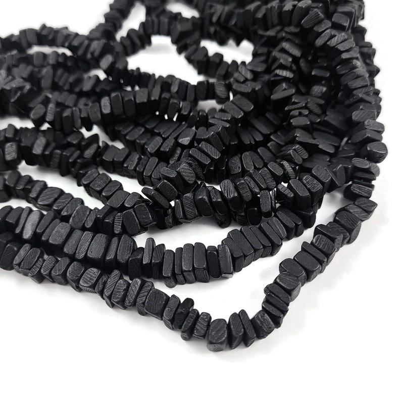 May include: Close-up of strands of black wooden beads. The beads are rectangular and have a slightly rough texture. The beads are strung together, creating a layered effect. The background is white.