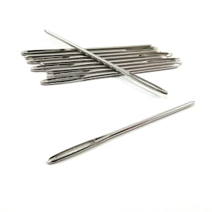 May include: A close-up of a pile of silver-coloured needles, with one needle lying separately on a white surface. The needles are long and thin, with a small eye at one end.