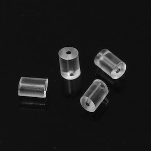 May include: Four transparent, cylindrical earring backs are displayed against a black backdrop. Each back has a central hole. The earring backs are made of a clear material, allowing light to pass through.