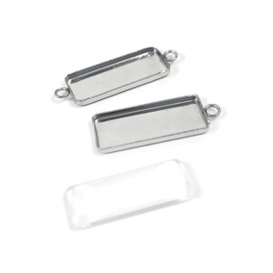May include: Three rectangular silver-coloured jewellery bezel trays with loop attachments, and a clear rectangular glass cabochon. The trays are empty and ready for use in jewellery making. The items are on a white background.