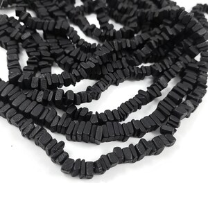 May include: Close-up of multiple strands of black, rectangular beads. The beads are strung together, creating a textured and organic appearance. The beads are a solid black color and appear to be made of a natural material.