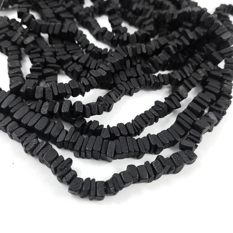May include: Close-up of multiple strands of black, rectangular beads. The beads are strung together, creating a textured and organic appearance. The beads are a solid black color and appear to be made of a natural material.