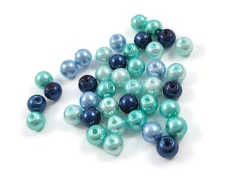 May include: A collection of assorted blue and teal beads scattered on a white surface. The beads vary in shades, including light blue, navy, and seafoam green. These round beads are likely for jewelry making or craft projects.