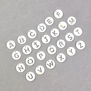 May include: A selection of small, circular, silver-coloured charms, each displaying a different letter of the alphabet. The letters are cut out of the metal, and each charm has a small hole at the top for threading. The charms are arranged on a grey surface.