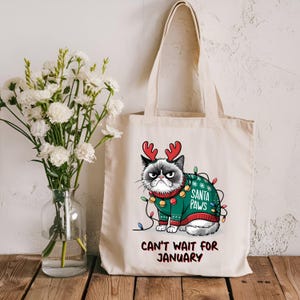 May include: A white canvas tote bag with a grumpy cat wearing a green Christmas sweater with red reindeer antlers and the text "Santa Paws" and "Can't Wait for January".