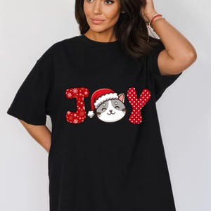 May include: Black t-shirt with a red, white, and gray graphic of the word "JOY" with a cat wearing a Santa hat in the middle of the word.