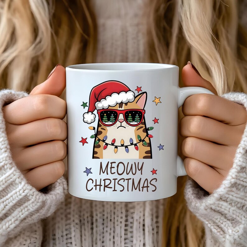 May include: White ceramic mug with a cartoon cat wearing a Santa hat and sunglasses. The cat is surrounded by Christmas lights and stars. The text "MEOWY CHRISTMAS" is printed on the mug.