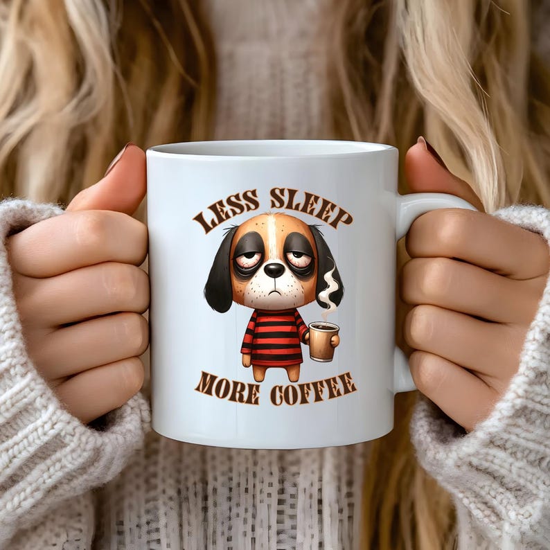 Op de afbeelding: Een witte keramische koffiemok met een cartoon-hond illustraties. De hond heeft bruin en wit haar, een rood-wit gestreept shirt en houdt een kopje koffie vast. De tekst op de mok luidt "Less Sleep More Coffee".