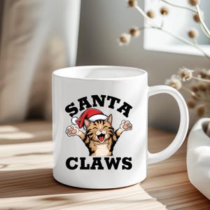 May include: White ceramic mug with a black and white illustration of a cat wearing a Santa hat. The text "SANTA CLAWS" is printed on the mug.