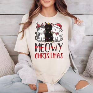 May include: A cream-colored t-shirt with a graphic of three cats wearing Christmas hats and a string of lights. The text "MEOWY CHRISTMAS" is printed below the cats.