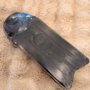 May include: A black plastic item, possibly a shoe insert, is packaged in clear plastic. The item has a curved shape with multiple sections. The packaging is on a fluffy, light brown surface.