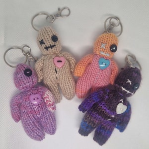May include: Four handmade crocheted voodoo dolls with keychains. Each doll has a different colour and design, including pink, purple, brown, and black. The dolls have stitched eyes and mouths, and each has a heart-shaped button on the chest.