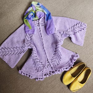 Puede incluir: Un cárdigan de ganchillo lila hecho a mano con detalles calados y mangas acampanadas. Una bufanda colorida con tonos azules, verdes y morados descansa sobre el cárdigan. Zapatos planos de cuero amarillos completan el conjunto.