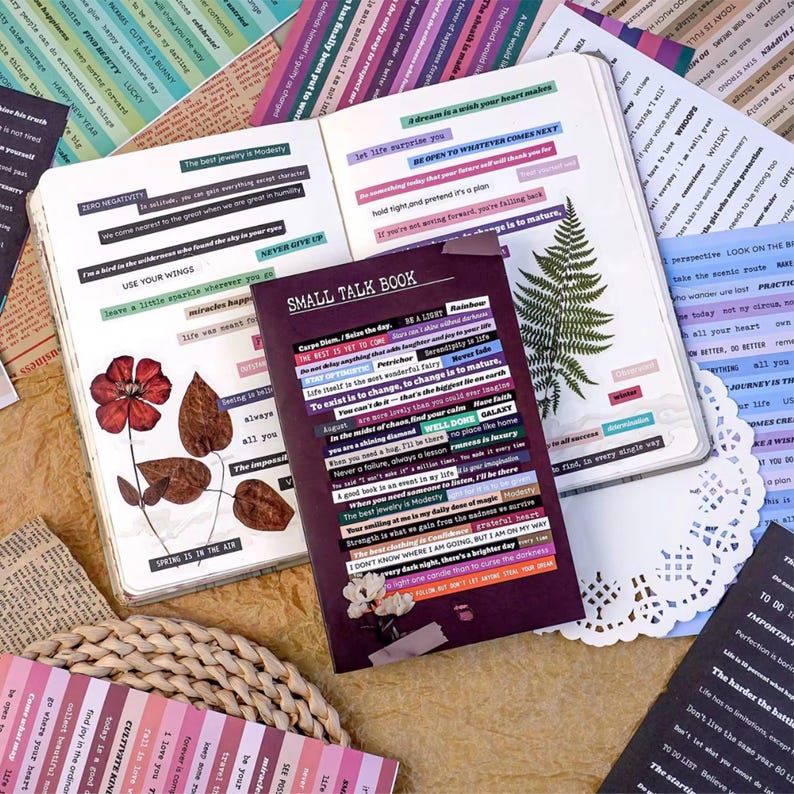 May include: A collection of stationery items, including a notebook titled "SMALL TALK BOOK", open journals, and strips of paper with printed text. The color palette includes shades of purple, pink, and white, with dried flowers and a fern leaf adding natural elements.