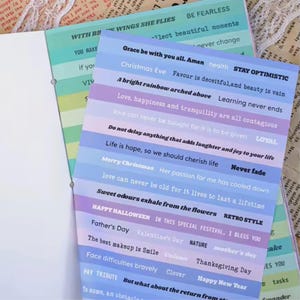May include: A close-up of decorative paper sheets with inspirational quotes and phrases in white font. The sheets have a pastel color scheme of blue, purple, and green stripes. The text includes phrases like "Stay Optimistic" and "Happy Halloween."