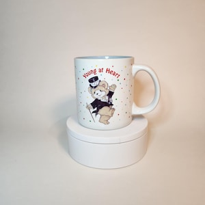 May include: White ceramic mug with a blue interior and a handle. The mug features a cartoon bear in a tuxedo and top hat, with the words "young at Heart" in red. Colorful confetti dots the mug's surface.