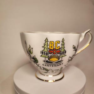 May include: White teacup with gold trim and a handle. The cup features a colourful emblem with the letters "BC", the years "1858" and "1958", and the word "Centenary". The cup has a floral design in black and green.