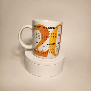 May include: White ceramic mug with a calendar design in orange and yellow. The mug displays months and dates. The mug has a white handle and is on a white surface. The calendar design includes the year 2019.