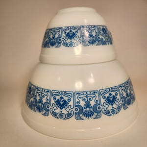 May include: Two white mixing bowls stacked. Each bowl features a blue decorative band with floral and heart-shaped designs. The bowls are made of a glossy material.