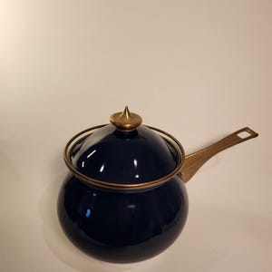 May include: A navy blue saucepan with a lid and a long handle. The pot has a rounded shape with a gold-coloured handle and lid accents. The lid has a pointed finial.