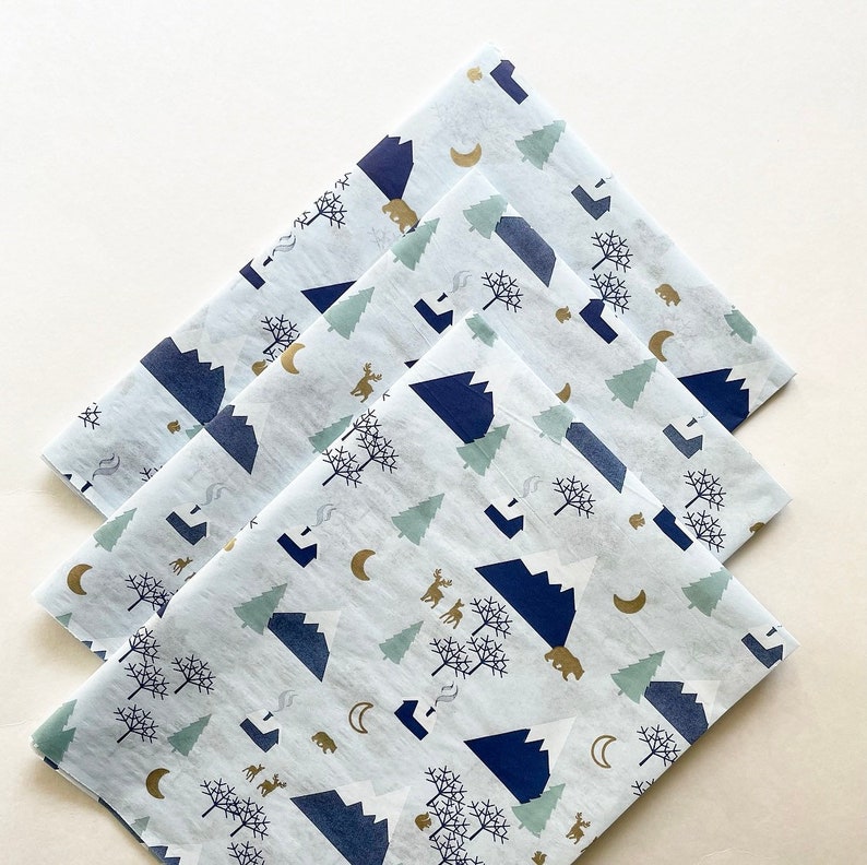 May include: Three sheets of white tissue paper with a blue and gold mountain and forest design. The design features mountains, trees, bears, deer, and moons.