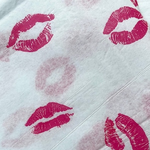 May include: Close-up of a white surface with multiple imprints of red lipstick lips. The lip prints vary in size and orientation, creating a pattern across the surface. The texture of the surface appears to be slightly wrinkled.