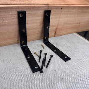 May include: Two black metal corner brackets with pre-drilled holes, alongside a collection of screws. The brackets are positioned near a wooden plank. The screws are in gold and black. Hardware for construction or woodworking.