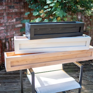 May include: Three wooden planter boxes stacked on top of each other. The top box is black, the middle box is white, and the bottom box is a natural wood color.