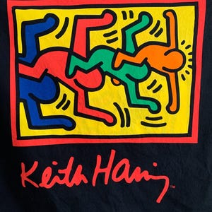 Op de afbeelding: Een zwart T-shirt met een kleurrijke afbeelding van drie figuren in een geel vierkant met een rode rand. De tekst "Keith Haring" is in rood onder de afbeelding gedrukt.