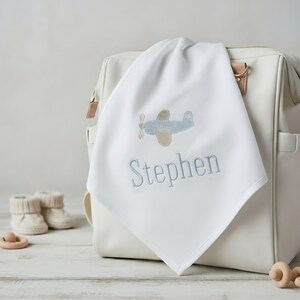 May include: A white baby blanket embroidered with a light blue airplane and the name "Stephen" in light blue thread. The blanket is draped over a white diaper bag. Baby booties and a wooden toy are in the background.