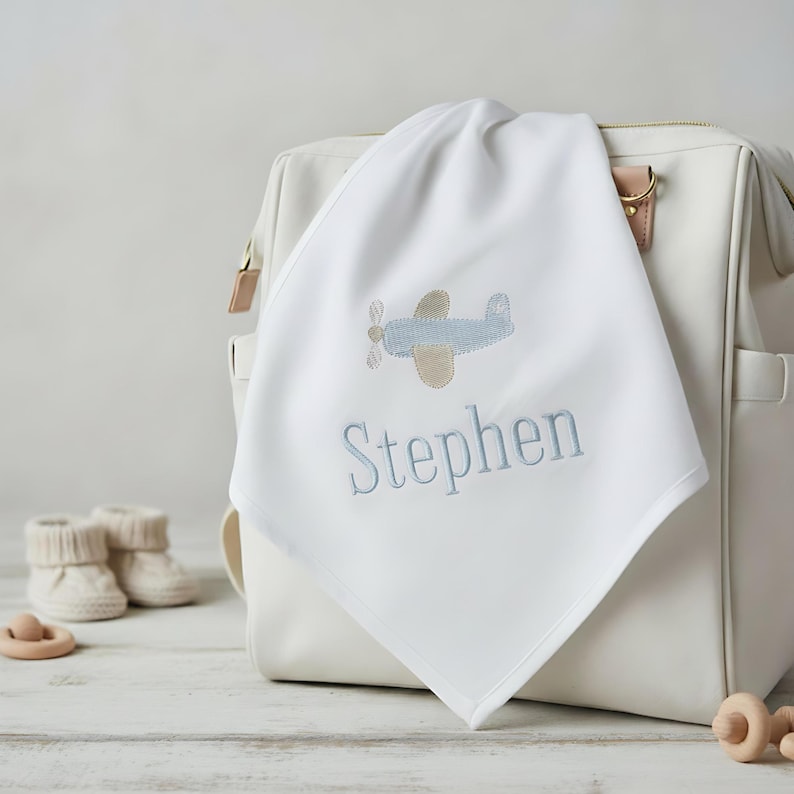 May include: A white baby blanket embroidered with a light blue airplane and the name "Stephen" in light blue thread. The blanket is draped over a white diaper bag. Baby booties and a wooden toy are in the background.