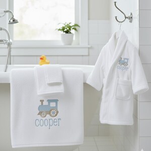 May include: White bath towel and washcloth set with a blue train design and the name "cooper" embroidered in blue. A matching white bathrobe with the same train design and name hangs on a hook. A yellow rubber duck sits on the edge of the tub.
