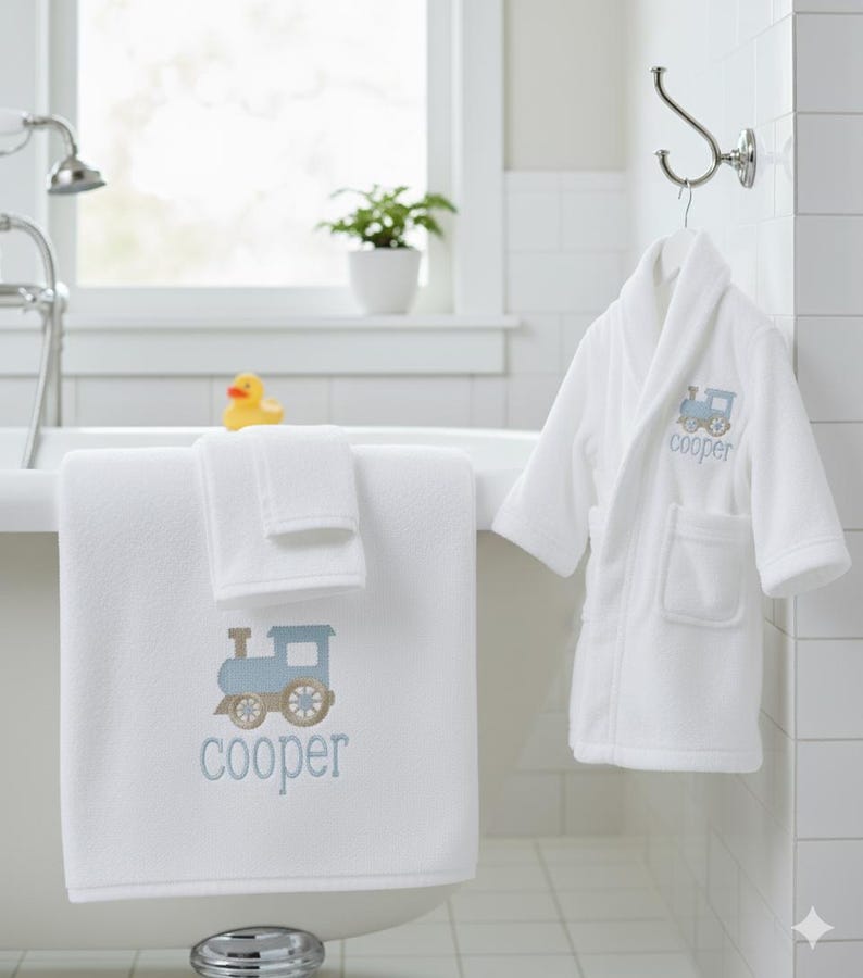 May include: White bath towel and washcloth set with a blue train design and the name "cooper" embroidered in blue. A matching white bathrobe with the same train design and name hangs on a hook. A yellow rubber duck sits on the edge of the tub.